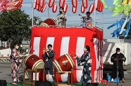 櫓の上で太鼓を叩く男性と太鼓を叩いている2人の浴衣を着た女性と横笛を吹いている男性の写真