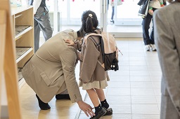 下駄箱前でくつを脱ごうとしている女児とそれを手助けしている親の写真