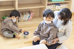 床に座っておもちゃで遊んでいる園児と先生の膝に座っている園児の写真