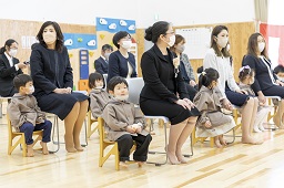 ホール内で椅子に座っている園児たちと親たちの写真