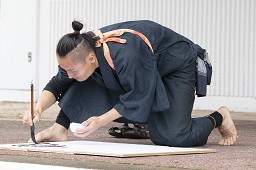 紺色の作務衣に橙色のたすき掛けをしている男性が舞台上で書道を披露している様子の写真