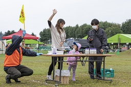 長机に置かれている空になった牛乳瓶と手を上げている女性と黄色い旗を上げているスタッフとタイマーを見ているスタッフの写真