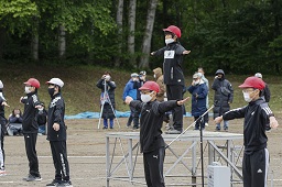 グラウンドの朝礼台の上の赤い帽子の小学生に合わせて準備体操をしている紅白の帽子をかぶった小学生たちの写真