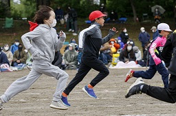 それぞれ赤い帽子や白い帽子をかぶっている4人の小学生たちがグラウンドで全力疾走している写真