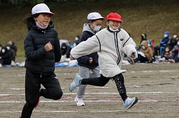 それぞれ赤い帽子や白い帽子をかぶっている3人の小学生がグラウンドで走っている様子の写真