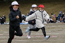 それぞれ赤い帽子や白い帽子をかぶっている3人の小学生がグラウンドで走っている様子の写真