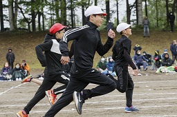 グラウンドで白い帽子の2人の小学生を追いかけている赤い帽子の小学生の競走をしている様子の写真