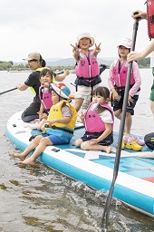 水に浮いているボードを漕いでいるスタッフと立ち上がってピースサインをしている女の子たちの写真