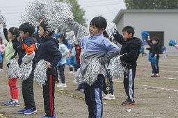 銀色のボンボンを両手に持ってグラウンドで二列になって踊っている小学生たちの写真