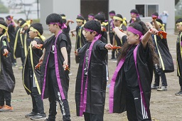 紫色のはちまきとハッピを着た小学生たちと黄色のはちまきとハッピを着た小学生たちがグラウンドで踊っている写真
