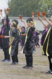 紫色のはちまきとハッピを着た小学生たちと黄色のはちまきとハッピを着た小学生たちがグラウンドで両手に楽器を持って踊っている写真