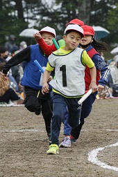 グラウンドでそれぞれバトンを持った白い帽子の小学生2人が赤い帽子の小学生2人より先を走っている様子の写真