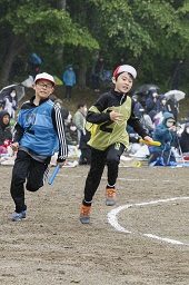 グラウンドでそれぞれバトンを持った赤い帽子の小学生が白い帽子の小学生の先を走っている様子の写真