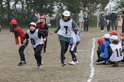 グラウンドで白い帽子と白いゼッケンを付けた小学生2人が赤い帽子と赤いゼッケンを付けた小学生2人より先に走る瞬間の写真