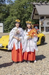 頭に飾りをつけて白い服と朱色の袴を着て紅葉した枝を持っている2人の女性の写真