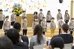 壇上で2列に並んで合唱をしている卒園児たちとそれを聞いている父母の写真