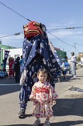 青い獅子舞の前で立っている赤い花柄の着物姿の子どもの写真