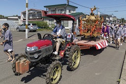 赤いトラクターの荷台に乗せられている金色の神輿と担ぎ手の男たちの写真