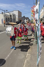 道路上で赤いハッピを着た子ども達が太鼓などの楽器を手に並んでいる様子の写真