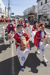 赤いハッピを着た子ども達が馬の頭を模した人形の手綱を握って行進している写真