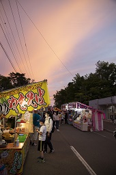 暮れなずむ空のもと道路沿いに並んでいるやきそばなどの屋台に集う人々の写真