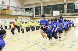 青いシャツを着た中学生たちと黄色いシャツを着た中学生たちが体育館で長縄跳びをしている写真