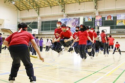 赤いシャツと赤いはちまきを付けた中学生たちが体育館で長縄跳びで飛んでいる瞬間の写真