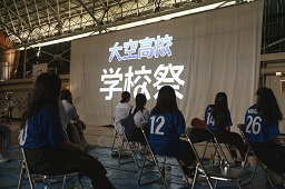 垂れ幕に大空高校学校祭とプロジェクターで映されているのを椅子に座って見ている高校生たちの写真