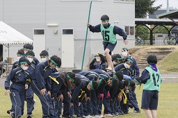 グラウンドで緑のはちまきとゼッケンを付けて緑の棒を持った男子中学生が緑のはちまきをした中学生たちのかがんだ背中の上を歩いている写真