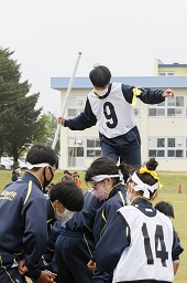 グラウンドで白のはちまきとゼッケンを付けて白い棒を持った男子中学生が白いはちまきをした中学生たちのかがんだ背中の上を歩いている写真