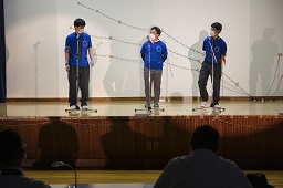 ステージ上で青いシャツを着た3人の男子高校生がマイクの前で話をしている写真