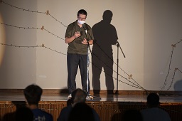 ステージ上でシャツを着た男性がマイクの前で話している写真