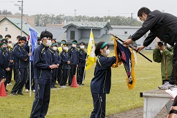 グラウンドで整列している中学生一同の前で朝礼台から優勝旗を受け取っている緑のはちまきをした女子中学生の写真