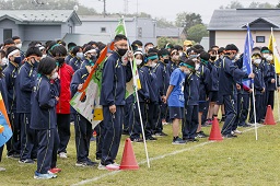グラウンドでそれぞれの色の旗の後ろに並んでいる中学生一同の写真