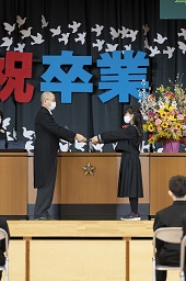 祝卒業と書かれた幕の前で校長先生から卒業証書を受け取っている女子卒業生の写真