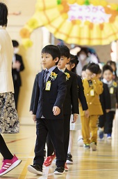 にゅうがくおめでとうと平仮名で書かれたゲートをくぐる新一年生たちが一列になって歩いている様子の写真