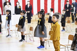 紅白幕が掛けられた体育館で椅子から立ち上がっている新一年生たちの写真