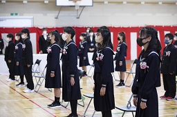 紅白幕が掛けられた体育館でバラのコサージュを制服の胸元につけている新一年生たちが起立している様子の写真