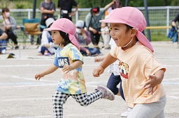 桃色の帽子をかぶった園児2人がグラウンドを走っている写真