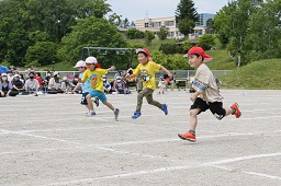 赤い帽子や白い帽子をかぶった4人の児童がグラウンドを走っている様子の写真