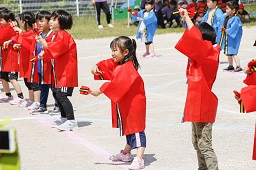 赤いハッピを着た園児たちがグラウンドで両手に持っている楽器を鳴らして踊っている様子の写真