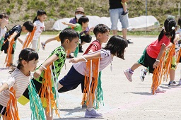 オレンジ色と緑色のビニールひもを腕につけている園児たちがグラウンドで創作ダンスを披露している写真
