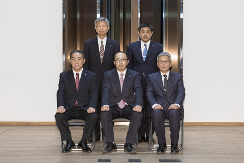 黒いスーツにピンク色のネクタイをしめた男性を真ん中に、前3名が座り後ろ2名が立ち正面を向いている写真