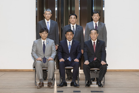 紺色のスーツに青色のネクタイをしめた男性を真ん中に、前3名が座り後ろ3名が立ち正面を向いている写真