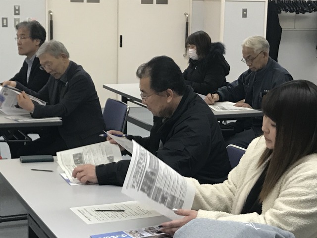 会議室で白い天板の長机に2名ずつで座り、資料を見ながら話を聞いている人たちの写真