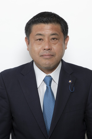 黒いスーツにブルーのほそいストライプのネクタイをしめた後藤忍さんが正面を向いている写真