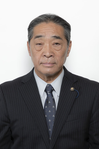 黒いストライプのスーツに薄いグレーのシャツ、紺色のネクタイをしめた松岡克美さんが正面を向いている写真