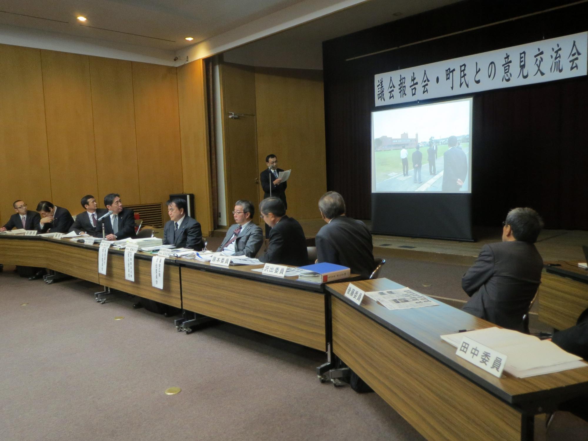 会議場で「議会報告会・町民との意見交流会」と書かれた垂れ幕の下に表示されるスライドを見る複数名の議会関係者の写真