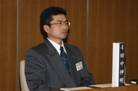 金縁の眼鏡をかけグレーのスーツ姿の田中伸明模擬議員が席に座り前を向いている写真