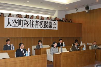 大空町移住者模擬議会の横断幕がさがった議事堂内の一階と中二階の席に複数の人が座り前方を向いている写真